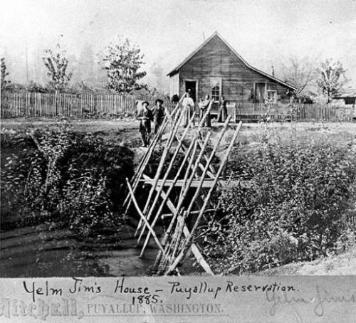 " House belonging to survivor of the Puget Sound Indian War, Yelm Jim [Wa-he-lut or Wahoolit], seen from across the water. Two men, three women pose in front of fenced house; in foreground is a large fish trap ." Source: http://digitalcollections.lib.washington.edu/cdm/ref/collection/loc/id/2092 